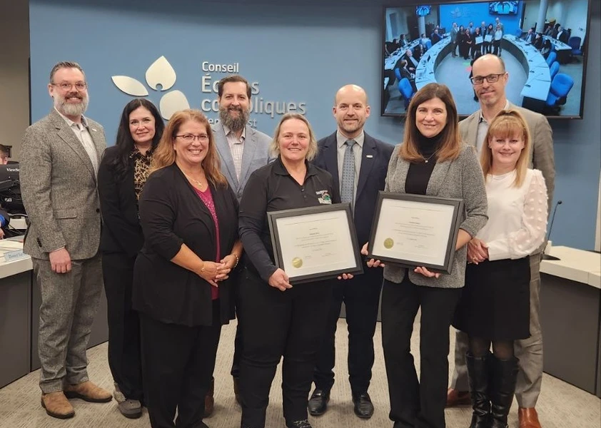 Groupe de huit adultes posant devant le mur du Conseil des écoles catholiques, deux femmes tenant des certificats encadrés lors d’une remise officielle.