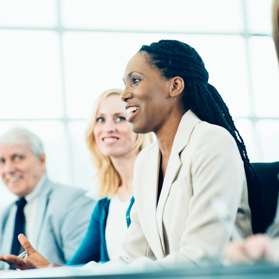 Groupe de professionnels en réunion dans un environnement lumineux. Une femme souriante en costume clair prend la parole, entourée de collègues attentifs et engagés.